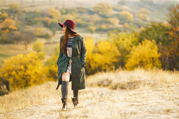 Beautiful young stylish girl in a coat walks in the autumn in the park. The girl is dressed in a green coat and a red hat. Beautiful evening. Autumn fashion. Lifestyle. High fashion portrait. 