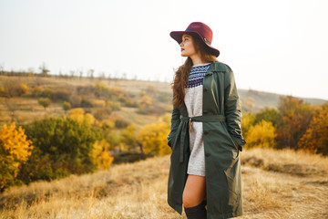 Beautiful young stylish girl in a coat walks in the autumn in the park. The girl is dressed in a green coat and a red hat. Beautiful evening. Autumn fashion. Lifestyle. High fashion portrait. 