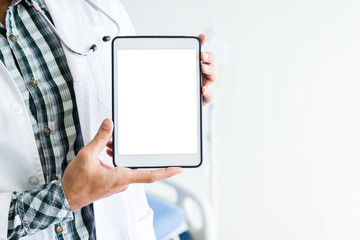 Close-up of man doctor therapeutic advising with positive emotions  holding up and showing digital tablet with a blank white screen and bed in hospital background,copy space