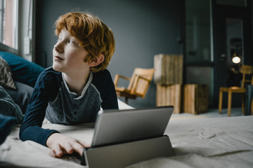 Redheaded boy lying on couch with digital tablet looking out of window