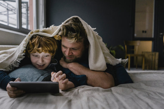 Father and son lying together under blanket looking at digital tablet