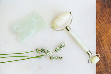 Gua sha and jade roller arranged with wildflowers / still life on white marble background with wood...