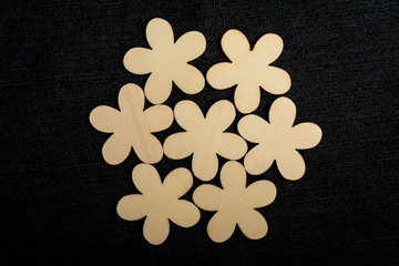Seven delicate light brown wooden flowers on dark grey textile material background, displayed centered, top view with space for text around, flat lay with laser cut wooden objects, selective focus