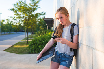 Naklejka na ściany i meble Depressed/Sad teen girl leaning against high school wall during sunset while wearing a backpack and holding binders/smartphone.