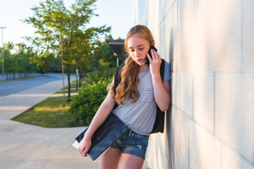 Depressed/Sad teen girl leaning against high school wall during sunset while wearing a backpack, holding binders, and talking on a smartphone.