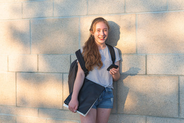 Teen girl leaning against high school wall during sunset while wearing a backpack and holding binders/smartphone.