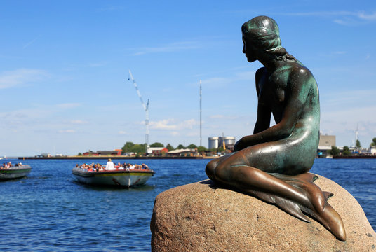 Copenhagen, Denmark - June 27, 2018: The Little Mermaid bronze statue by Edvard Eriksen, with sightseeing boats in the background.
