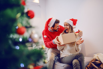 Amazed cute Caucasian blonde woman sitting on sofa in living room and receiving gift from her boyfriend. Both having santa hats on heads. In foreground is christmas tree. Living room interior. - Powered by Adobe