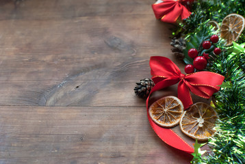 Fototapeta premium Christmas background with decorative composition of green tinsel, dried oranges, red berres, pine cone, gift with red bow and stars on wooden board. Happy new year.