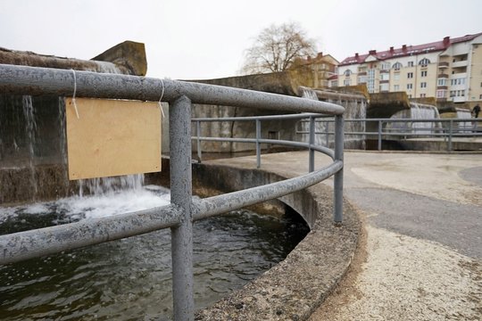 concrete structure in the form of a dam with a metal fence of round pipes with a wooden sign for the inscription, an artificial pond with a waterfall and foaming water