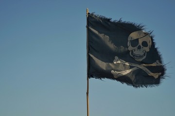 A pirate flag is developing in the sky. The flag is black with a skull. Pirate flag on blue sky background