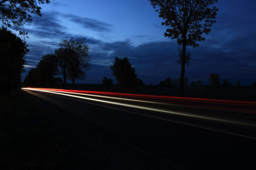 Światła samochodów w nocy na drodze między polami o zmierzchu. - obrazy, fototapety, plakaty