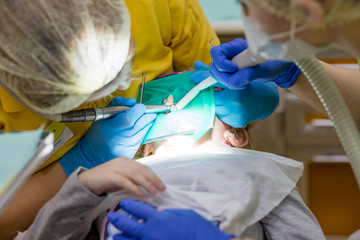 Dentist performing dental filling procedure to preteen girl in pediatric dental clinic. Doctor removing caries using high-speed dental drill. Calm child is sitting in a dental chair holding mouth open
