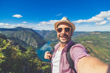 Fashionable tourist taking photo of himself while standing on hill above canyon and mountains.