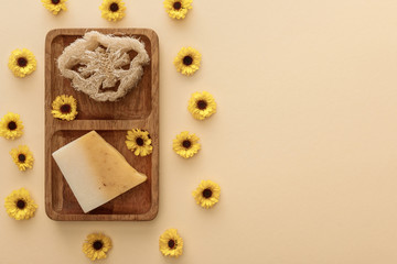 Fototapeta na wymiar top view of brown wooden soapdish with loofah and piece of soap on beige background with flowers