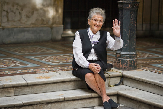 Portrait of a senior adult woman sitting and waving on a step in a foyer.