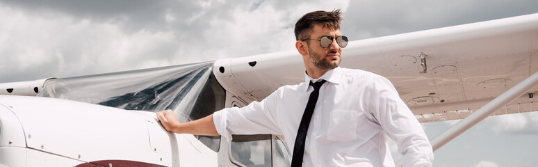 panoramic shot of confident pilot in sunglasses standing near plane - obrazy, fototapety, plakaty