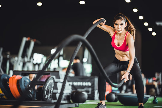 Beautiful Asian woman exercise on battling ropes training equipment in indoor fitness gym. Sport recreational activity, people workout, or healthy lifestyle concept