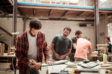 Man looking at male carpenter sanding wood at workbench with colleagues working in background