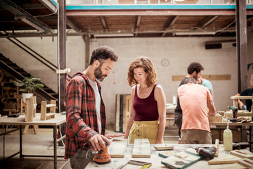 Smiling female carpenter looking at male colleague holding sander with coworkers working in background