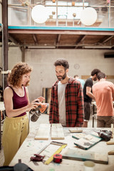 Smiling carpenter looking at female colleague holding sander while standing in workshop