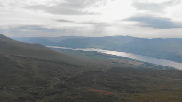 An aerial forward footage of a grassy mountain plateau with a huge lake and mountain range in the background under a cloudy sky	