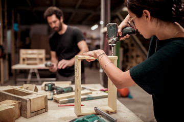 Female carpenter drilling wooden frame while working at workshop with colleague in background