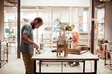 Carpenter making wooden object at workbench with colleague working in background