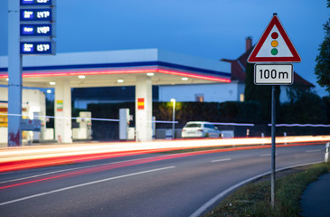 A triangular red white warning sign that shows that there is a traffic light 100 meters away. Opposite in the background a lit petrol station out of focus with beautiful bokeh.