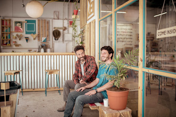 Cheerful male colleagues talking sitting on bench in workshop