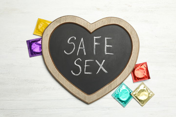 Colorful condoms and heart shaped blackboard with words SAFE SEX on white wooden background, flat lay