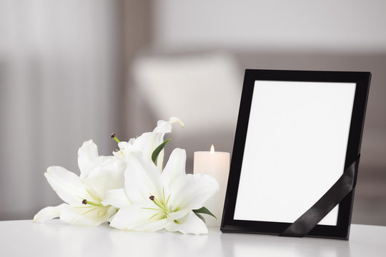 Funeral photo frame with black ribbon and lilies on white table indoors. Space for design