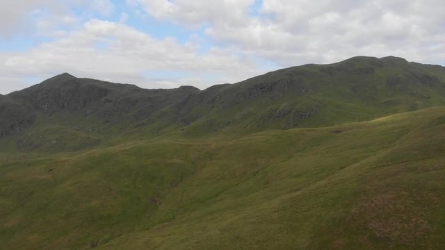 An aerial forward footage of a grassy mountain slope and rocky summit under a blue sky and white clouds