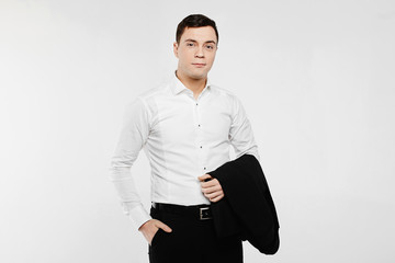 Obraz na płótnie Canvas Young and handsome businessman in the white shirt and dark pants holding blazer on his hand and posing over white background, isolate. Copy space from the left and right sides.