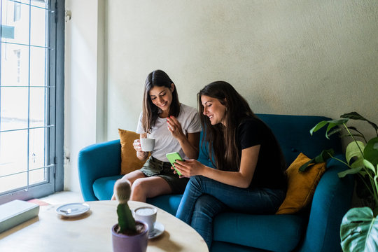 Two young female, friends sitting on a couch in a cafe sharing cell phone