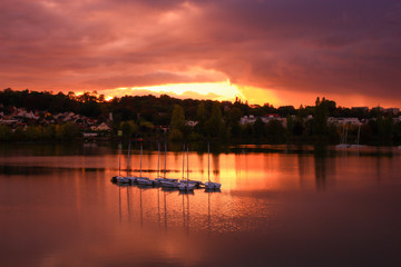 Group of sailboats on a lake at sunset. Houses and forest in the background. Colored dramatic sky and clouds. 