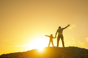 Father with a baby girl on top of the mountain, raising his hands up, playing outdoors on a sunset background. Happy loving family