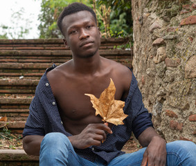 Hombre joven negro de Senegal sentado, con una hoja de otoño en las manos. - obrazy, fototapety, plakaty