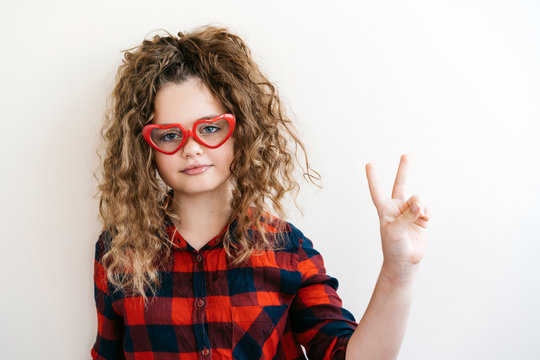 Cute curly long-haired blonde teen girl looks over funny heart-shaped glasses red color. The teenager smiles a beautiful snow-white smile. The concept of interest and good vision. Victory sign