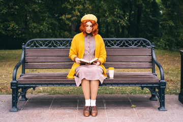 A beautiful red-haired girl with a curly hairstyle is sitting on a park bench and reading a book. A bamboo cup with hot coffee stands nearby. Fashionable autumn clothes in orange mustard tones