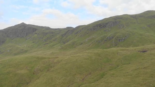 An aerial backward footage of a grassy mountain slope and rocky summit under a blue sky and white clouds