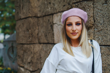 POrtrait of young blonde woman wearing white dress and beret standing in front of brown concrete stone wall in autumn day