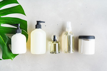 Cosmetic set of blank label bottles for mockup packaging of skincare product cream, serum, oil, shampoo, conditioner, perfume on grey background with green leaves. Natural beauty product concept.