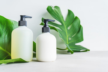 Cosmetic set of two blank label bottles for mockup packaging of skincare product cream, shampoo, conditioner on grey background with green leaves. Natural beauty product concept.