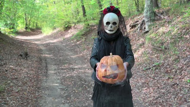 Woman close on with creepy skull mask in the autumn woods, holding a carved Halloween smoky pumpkin.  On background path with  fall leaves. Concept about Halloween holidays and Day of the dead.