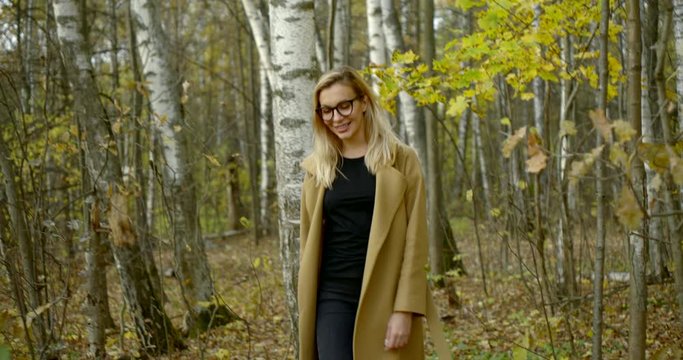 a young smiling blonde girl in glasses and a coat, she walks through the autumn forest, against the background of birches. She's wearing ripped black jeans.