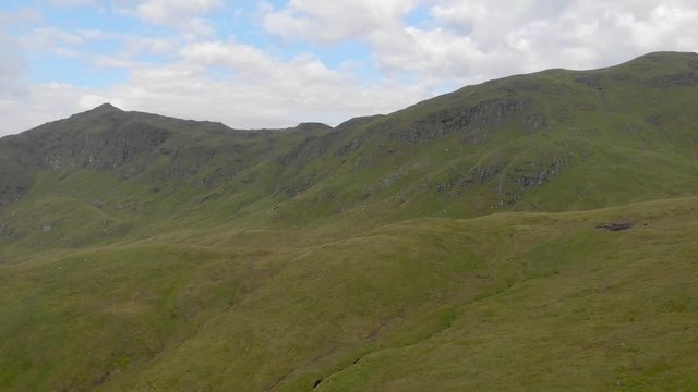 An aerial backward footage of a grassy mountain slope and rocky summit under a blue sky and white clouds
