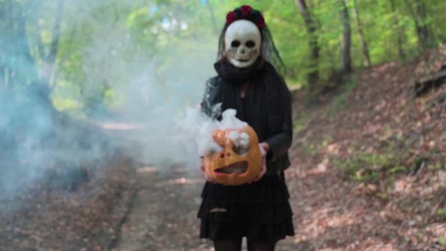 Woman  on defocus with creepy mask in the autumn woods, holding a carved Halloween smoky pumpkin. Concept about Halloween holidays.