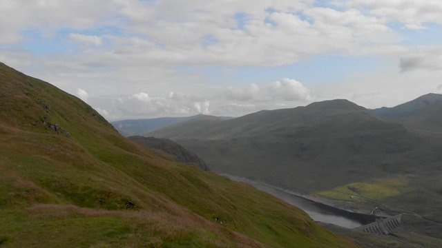 An aerial forward going down low altitude passing close to a pilot drone on a grassy summit with a lake, dam and mountain range in the background under a majestic blue sky and white clouds