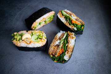Onigirazu with tempeh, spinach, carrot, cucumber and sesame sauce with other onigirazu filled with broccoli, cashews, cilantro and curry sauce on concrete background. Japanese food.